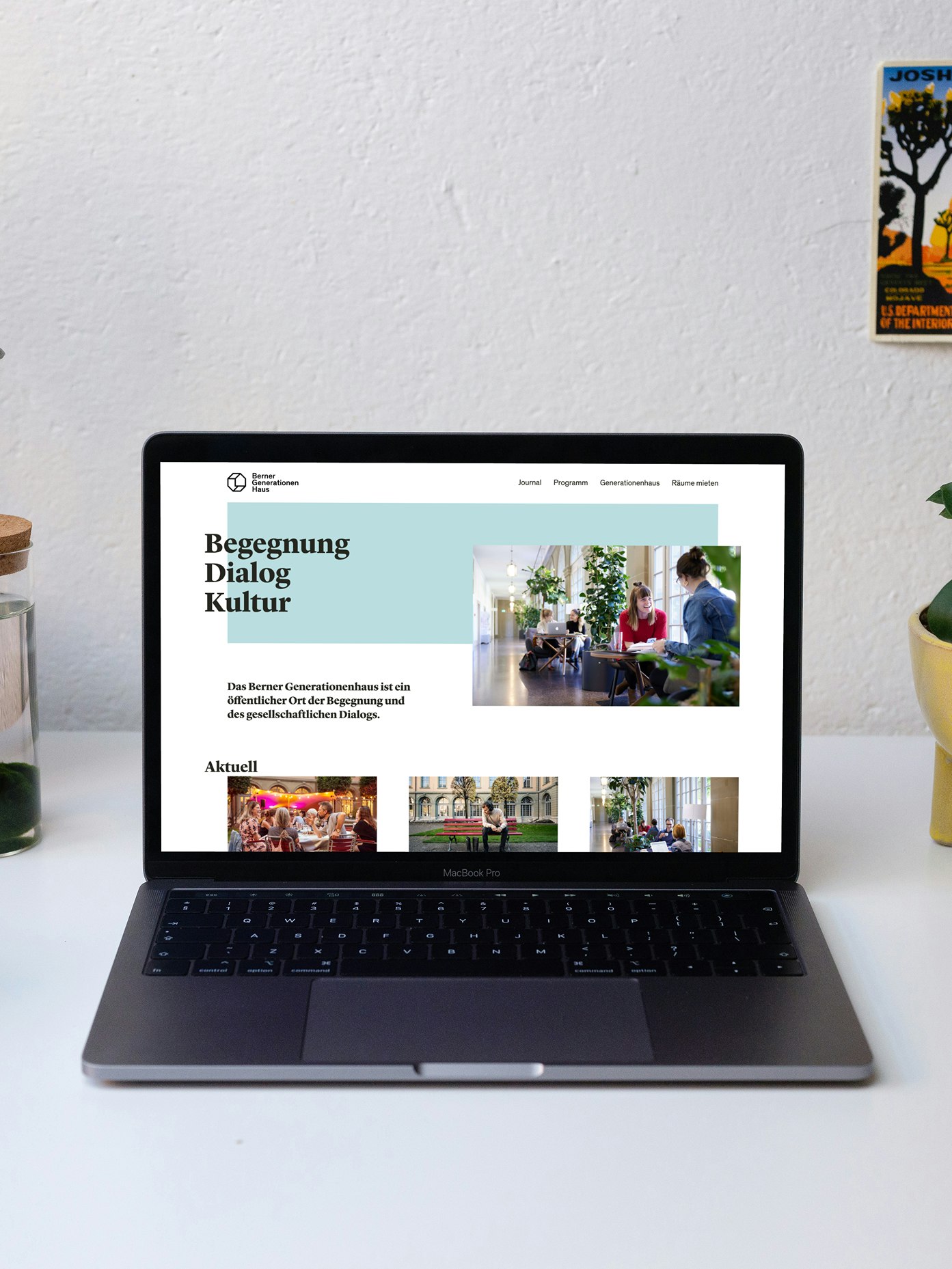 A laptop displaying a webpage on a desk with plants, a water bottle, and postcards in the background.