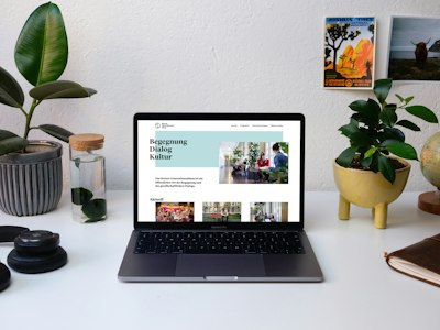 A laptop displaying a webpage on a desk with plants, a water bottle, and postcards in the background.