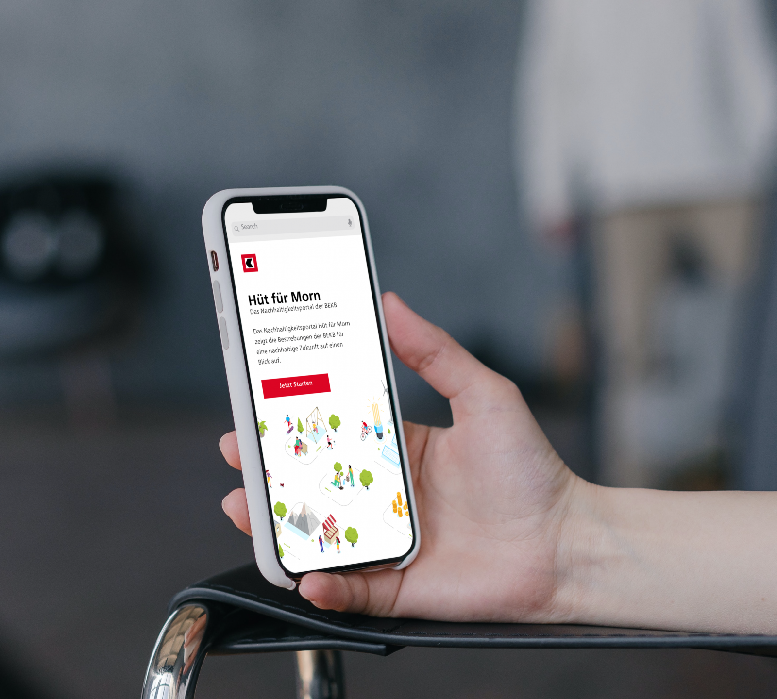 A hand holds a smartphone displaying a website titled "Hit für Morn" in German, featuring a colorful illustration of people and buildings, with a red button labeled "Jetzt Termin.