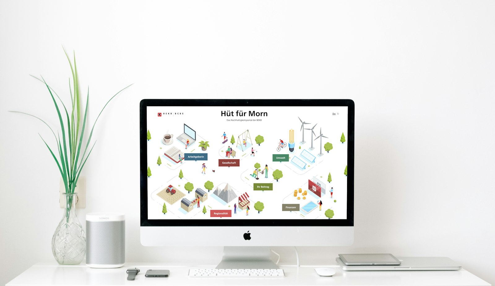 Computer on desk showing a webpage with an isometric illustration of people, buildings, and wind turbines. Nearby are a plant, speaker, phone, and keyboard.