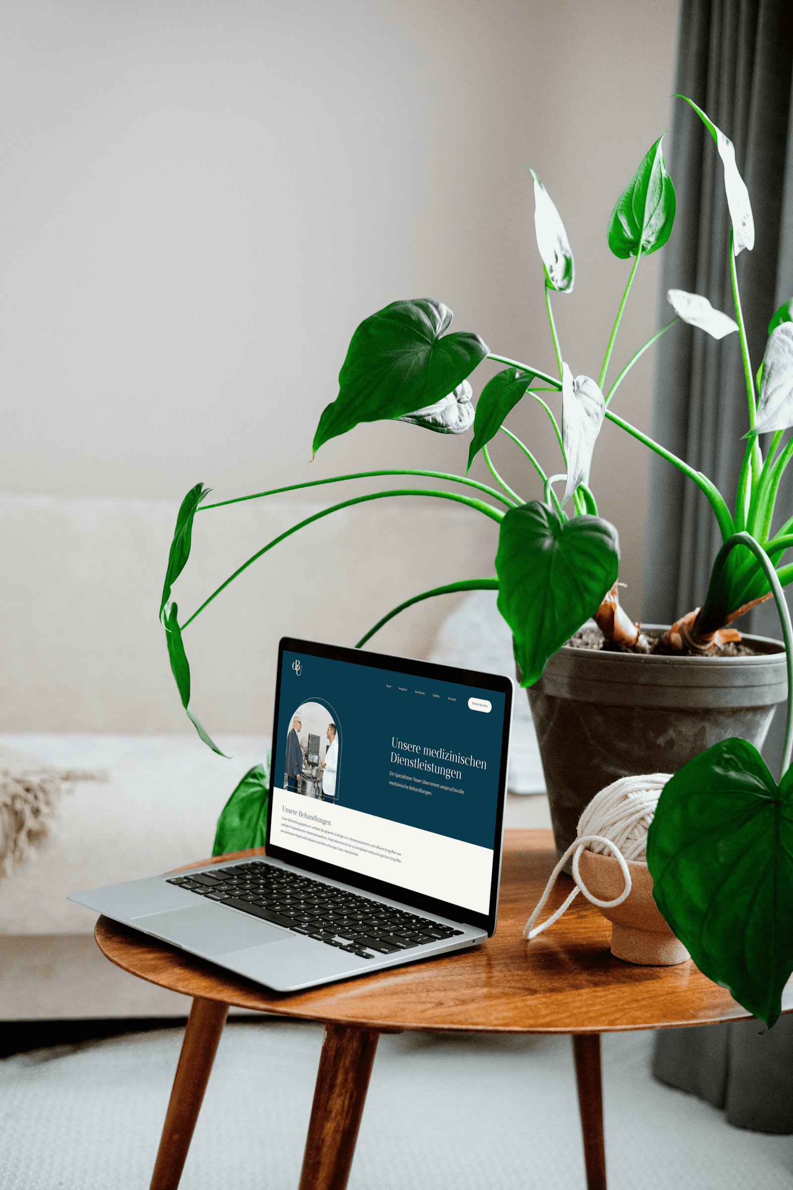 A laptop displaying a website is on a wooden table next to a large potted plant in a cozy living room setting.