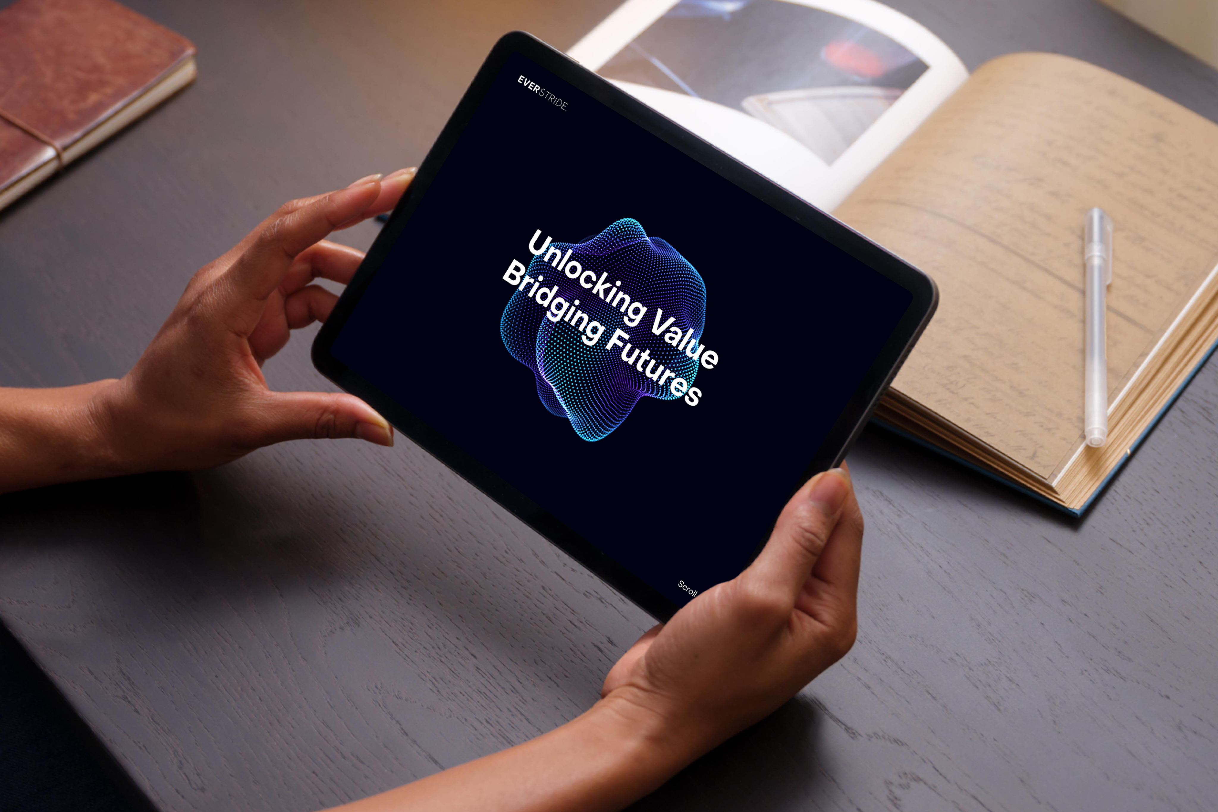 Person holding a tablet displaying the text "Unlocking Value Bridging Futures" with a blue graphic, on a dark table next to an open notebook and pen.
