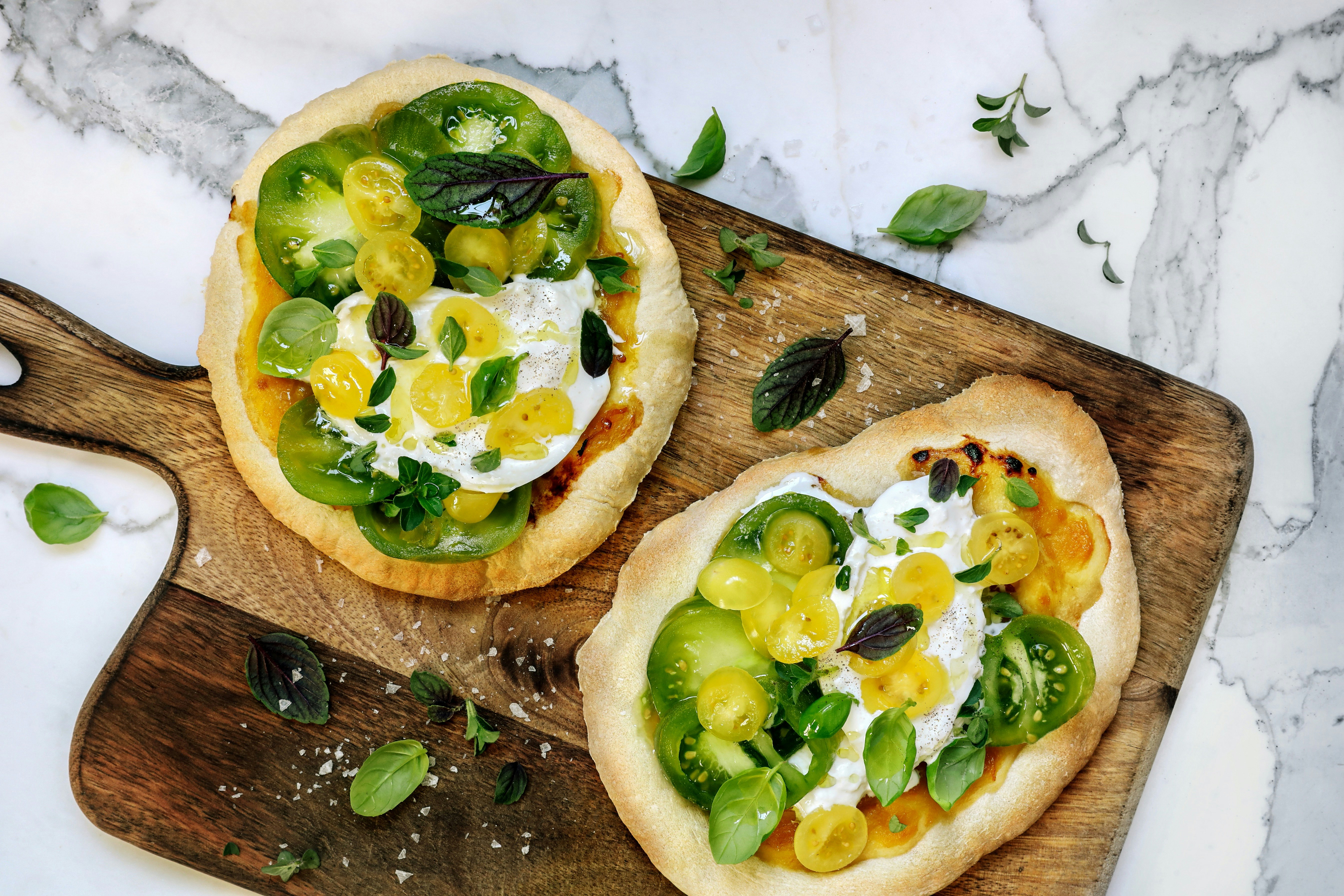 Two flatbread pizzas topped with green and yellow tomatoes, mozzarella, and fresh basil on a wooden board with marble background.