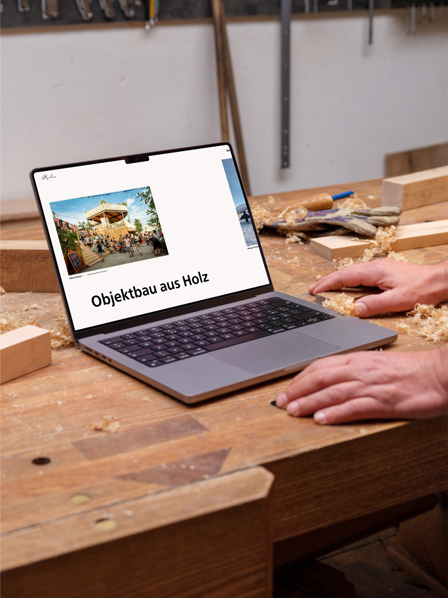 A person works at a wooden workbench with a laptop showing "Objektbau aus Holz," surrounded by wood shavings and tools.