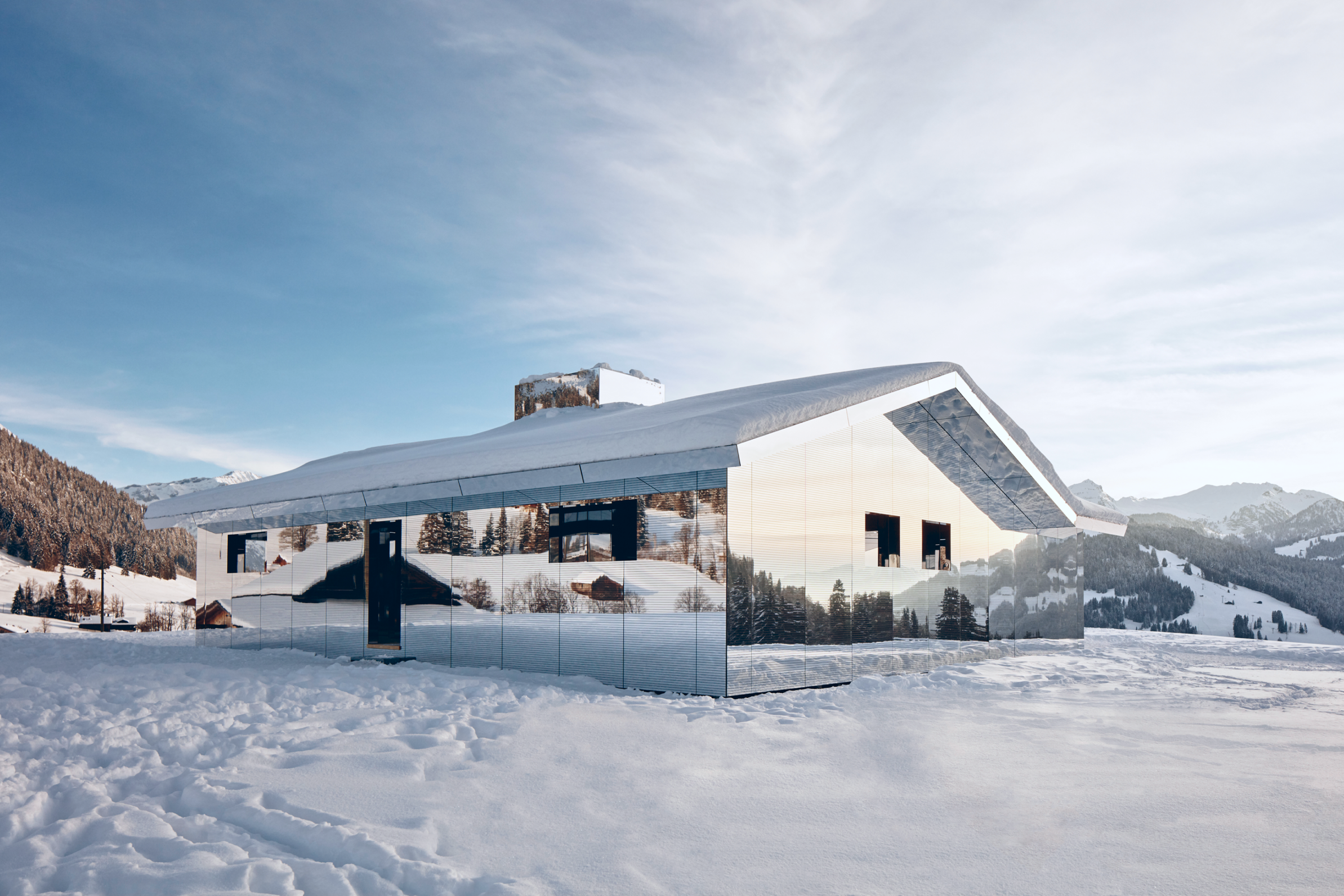 A house with mirrored walls reflects snowy mountains and trees, set in a winter landscape under a clear sky.