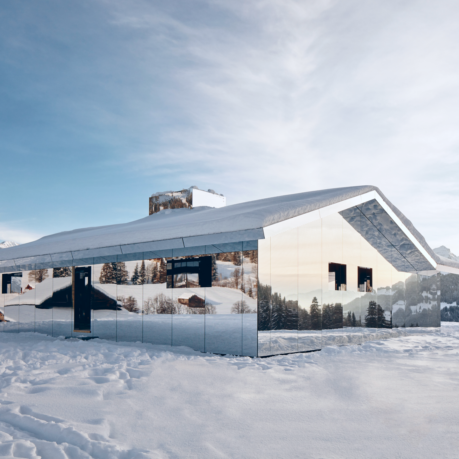 A house with mirrored walls reflects snowy mountains and trees, set in a winter landscape under a clear sky.
