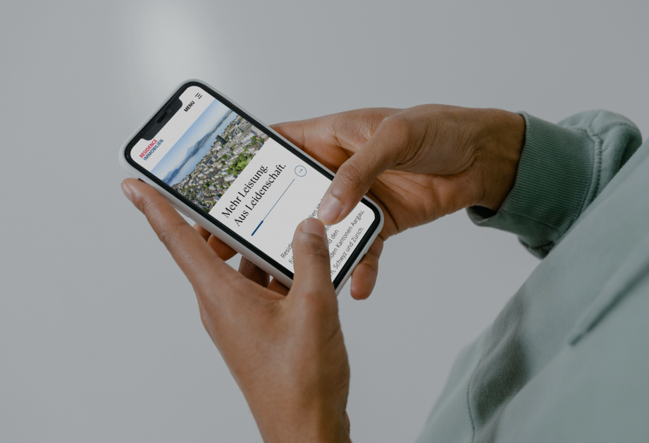 A person holds a smartphone displaying a webpage titled "More Learning, Less Judgement." The screen shows a photo of a landscape and text. The phone is held with both hands against a gray background.