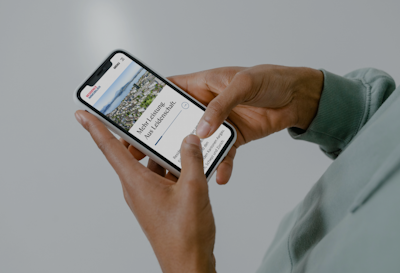 A person holds a smartphone displaying a webpage titled "More Learning, Less Judgement." The screen shows a photo of a landscape and text. The phone is held with both hands against a gray background.
