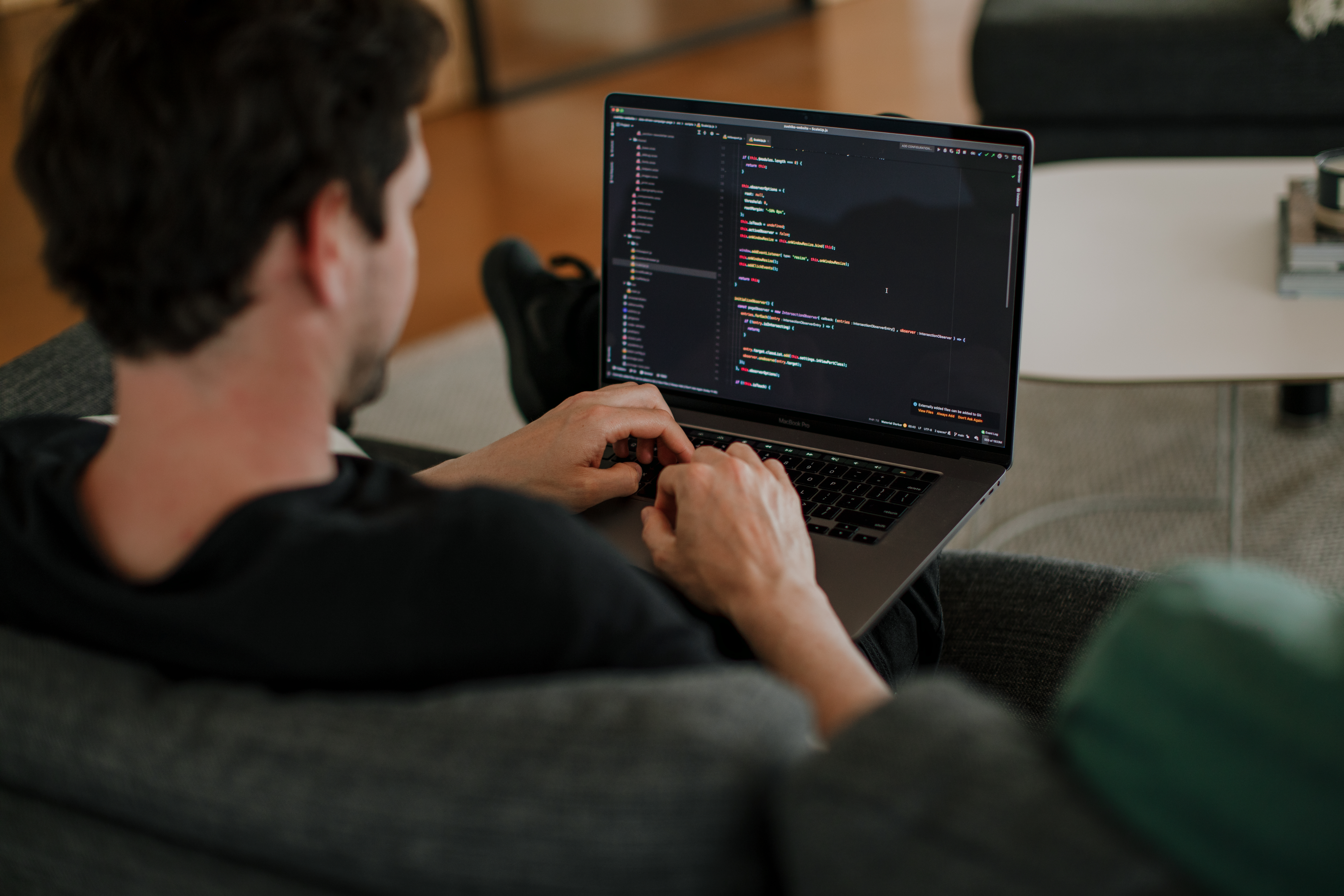 Eine Person sitzt auf einem grauen Sofa und tippt auf einem Laptop, auf dem in einem Texteditor bunter Code angezeigt wird. Die Umgebung scheint ein gemütlicher Raum mit sanfter Beleuchtung und minimalistischem Dekor zu sein.