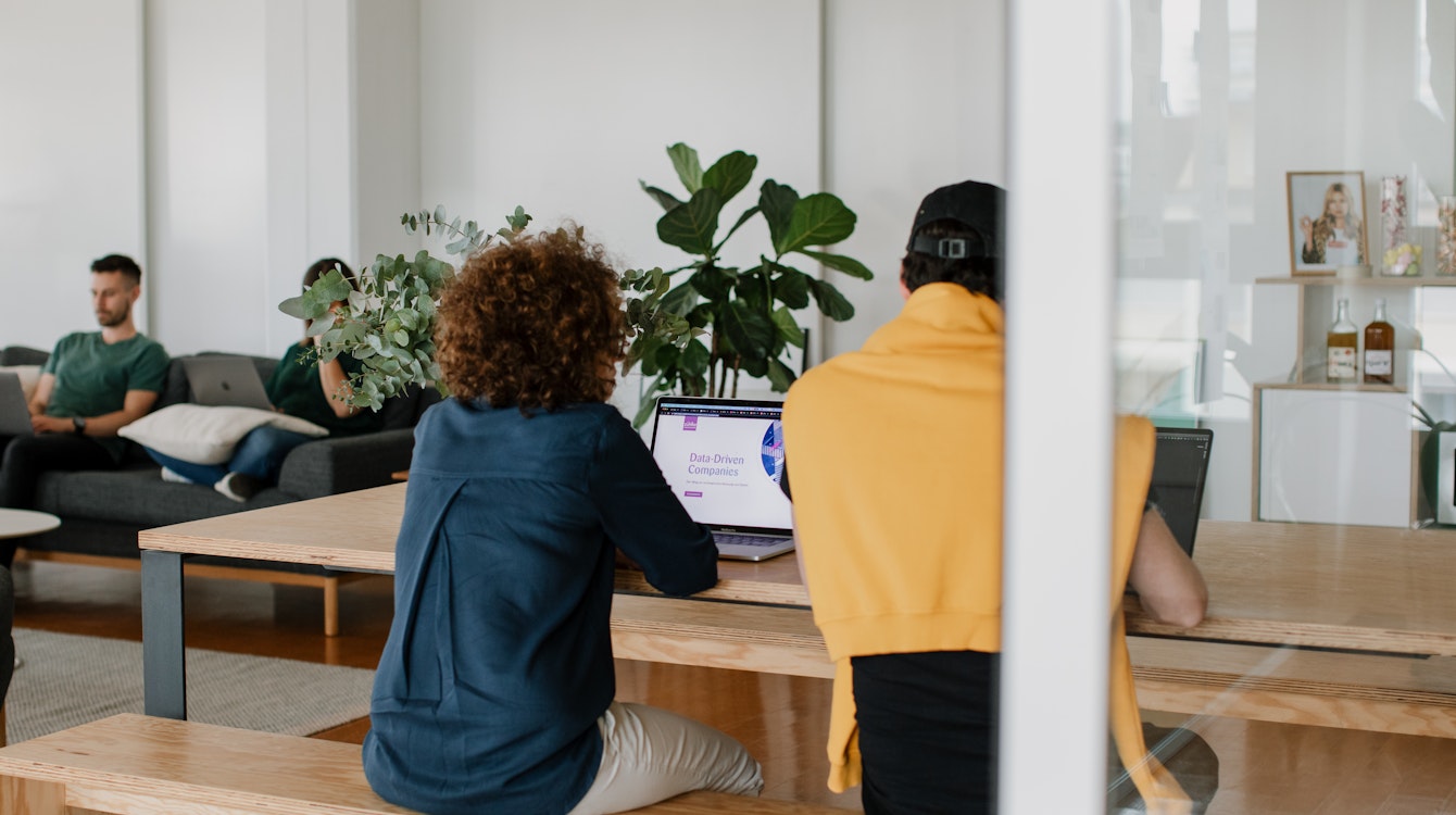Zwei Personen sitzen an einem Holztisch und arbeiten an Laptops in einem Büro mit großen Pflanzen. Auf dem Bildschirm wird eine Präsentationsfolie angezeigt. Im Hintergrund sitzt ein Mann auf einem Sofa neben einem Bücherregal mit Dekorationsgegenständen.