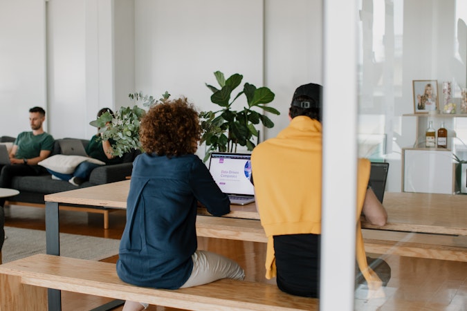 Zwei Personen sitzen an einem Holztisch und arbeiten an Laptops in einem Büro mit großen Pflanzen. Auf dem Bildschirm wird eine Präsentationsfolie angezeigt. Im Hintergrund sitzt ein Mann auf einem Sofa neben einem Bücherregal mit Dekorationsgegenständen.