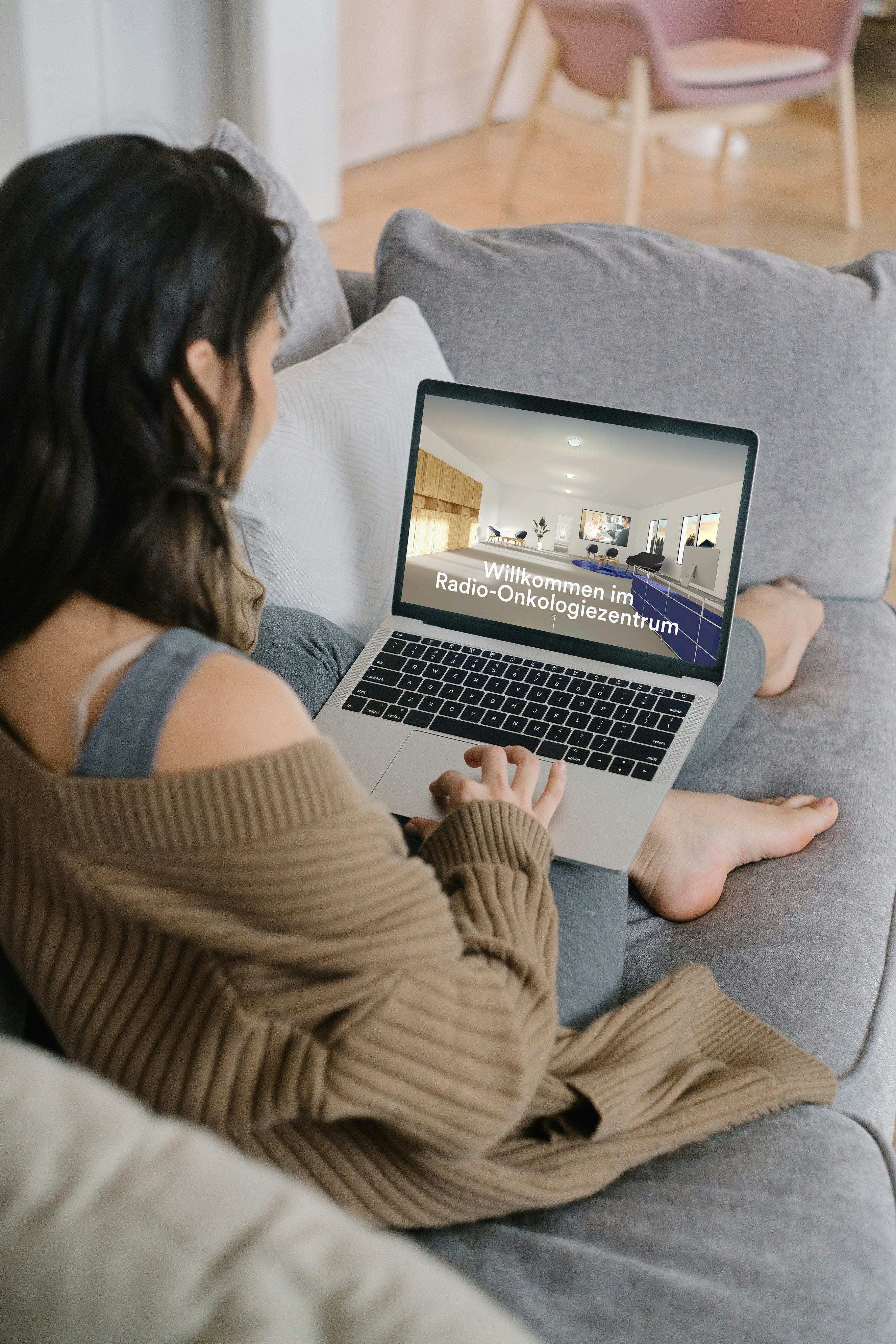 Eine Frau sitzt auf einer Couch und benutzt einen Laptop. Auf dem Bildschirm erscheint ein Bild einer medizinischen Einrichtung mit deutschem Text: Willkommen im Radio-Onkologiezentrum, was soviel bedeutet wie: Willkommen im Radio-Onkologiezentrum.