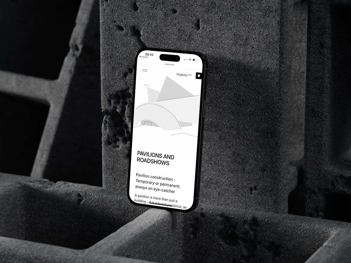Ein Smartphone, das eine Website über Pavillons und Roadshows anzeigt, steht aufrecht vor einem Stapel rauer, dunkler Betonblöcke. Der Bildschirm zeigt minimalistische Grafiken und schwarzen Text auf weißem Hintergrund.
