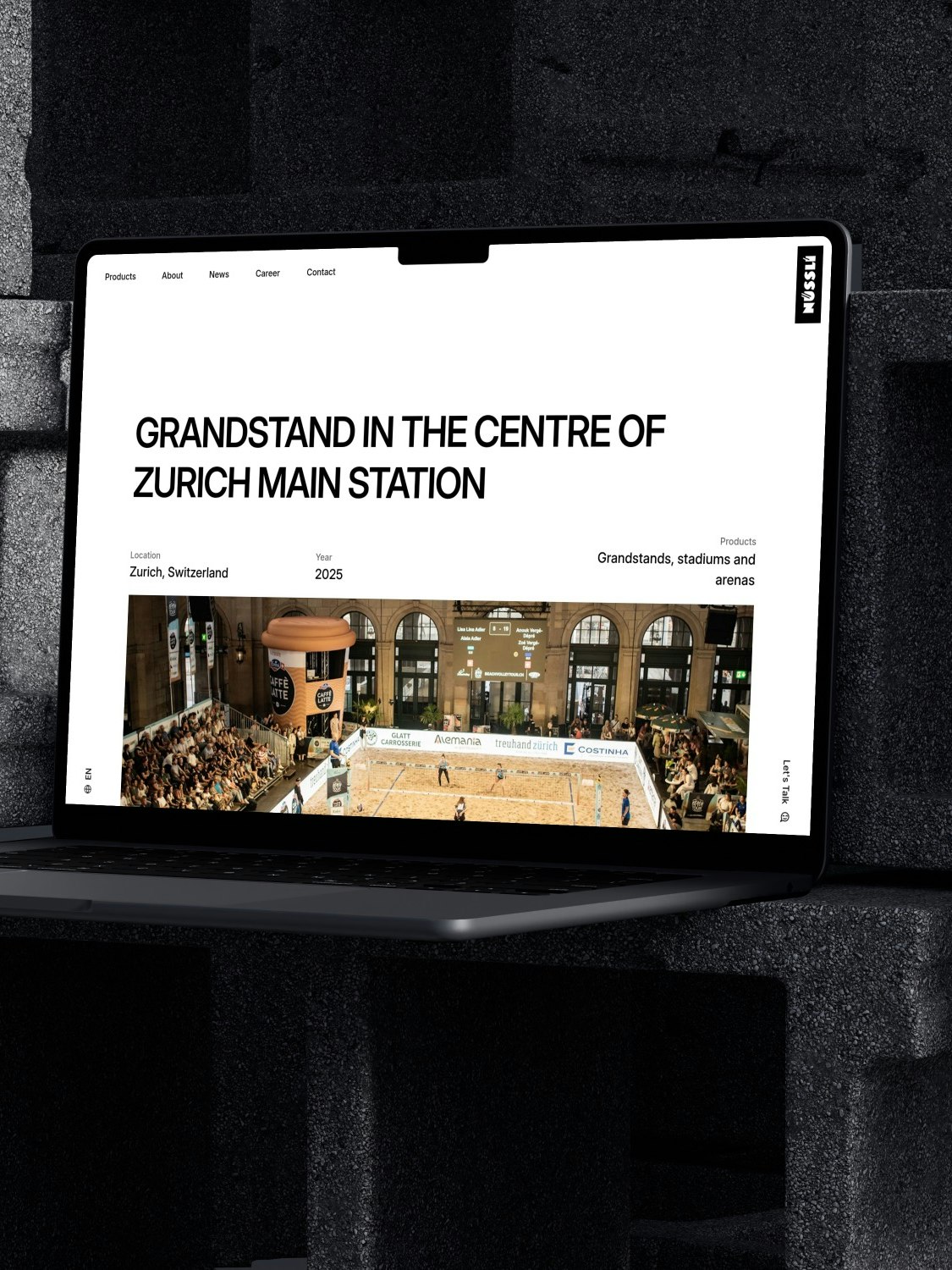 Ein Laptop ruht auf gestapelten Betonblöcken und zeigt eine Website mit der Überschrift Tribüne im Zentrum des Zürcher Hauptbahnhofs über einem Foto einer Sportveranstaltung in einer großen Halle.