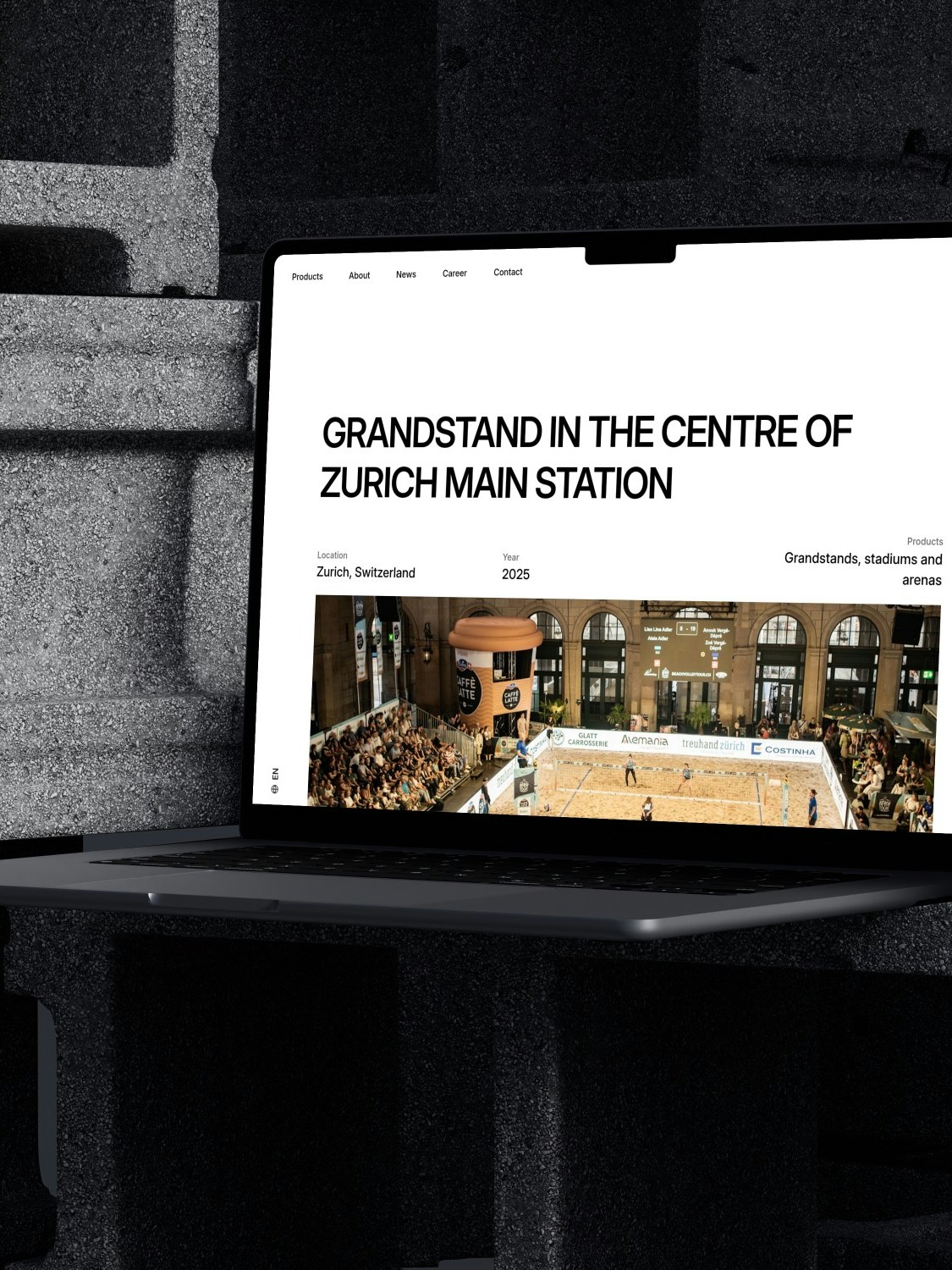 Ein Laptop ruht auf gestapelten Betonblöcken und zeigt eine Website mit der Überschrift Tribüne im Zentrum des Zürcher Hauptbahnhofs über einem Foto einer Sportveranstaltung in einer großen Halle.
