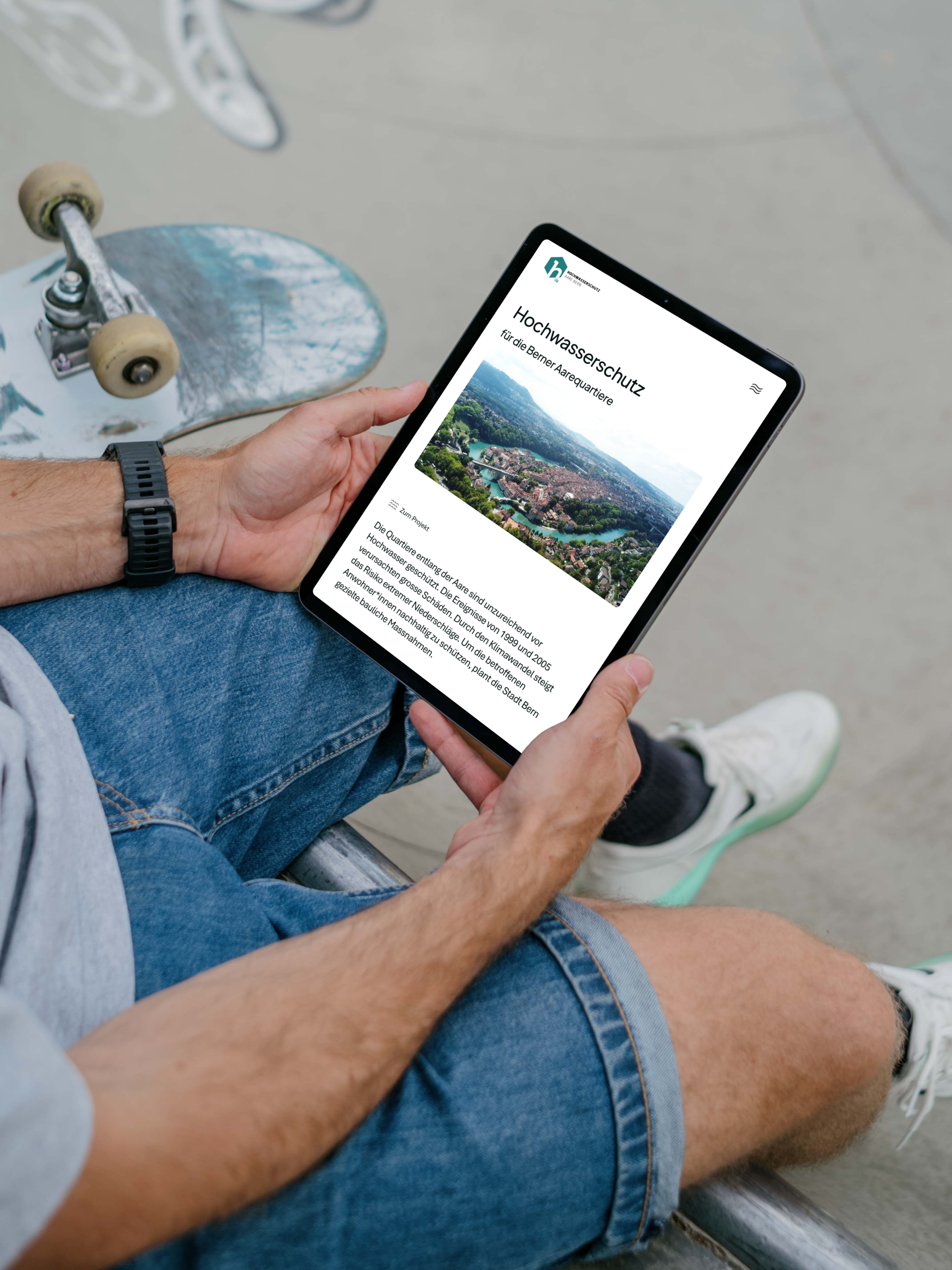 Eine Person, die mit einem Skateboard im Freien sitzt, hält ein Tablet in der Hand, auf dem ein deutscher Artikel über Hochwasserschutz zu sehen ist, der ein Luftbild einer Stadt und der umliegenden Grünflächen zeigt.
