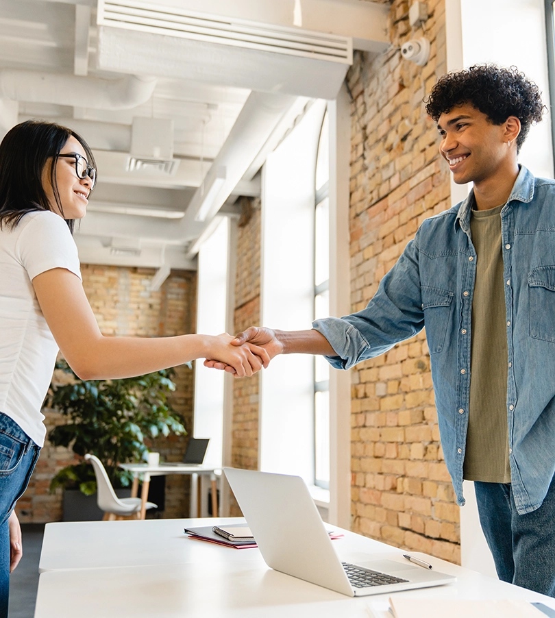 Two people shaking hands over a desk with a laptop on
