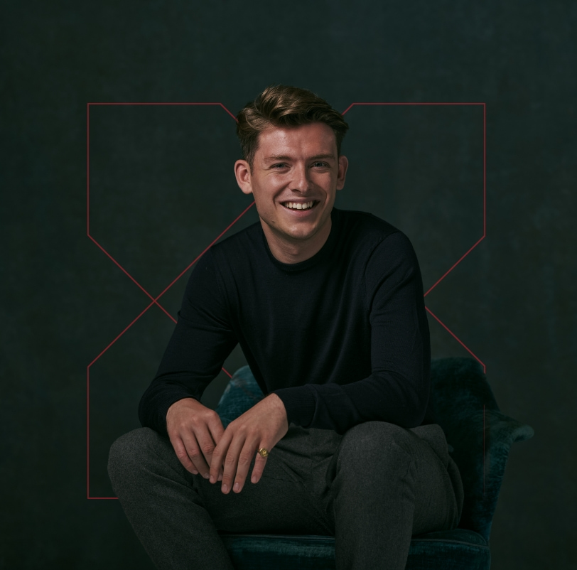 Headshot image of a man in black jumper sat on a velour chair. The backdrop is grey and moody.