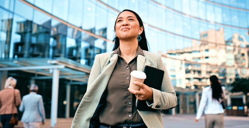 miling businesswoman holding coffee and documents while walking outside a modern office building