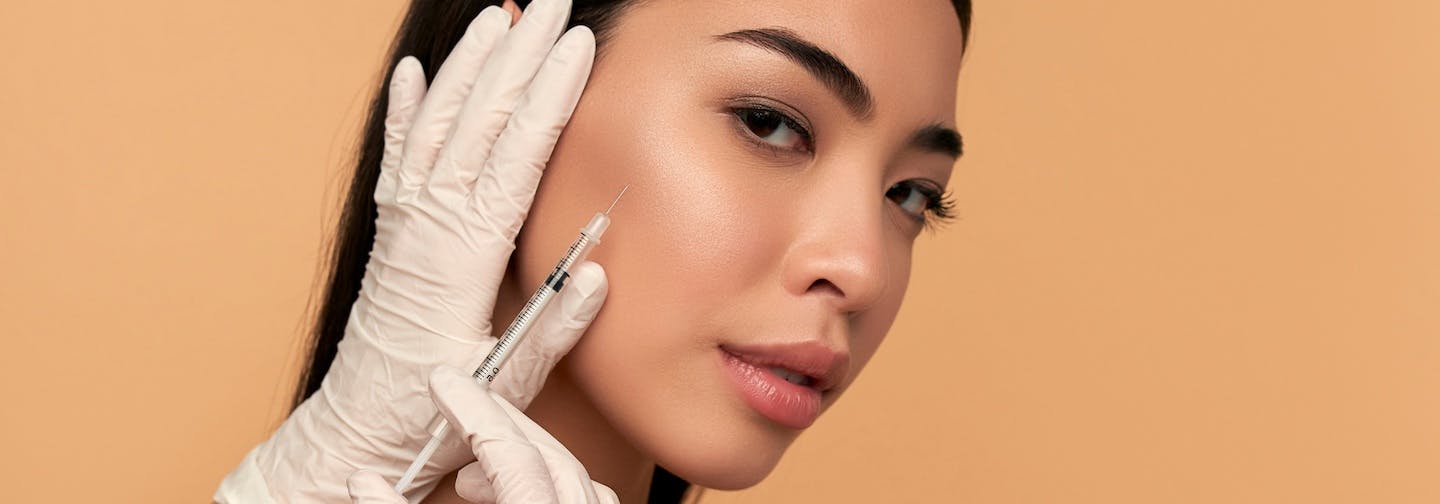 gloved hand holds dermal filler or neuromodulator injections needle near face