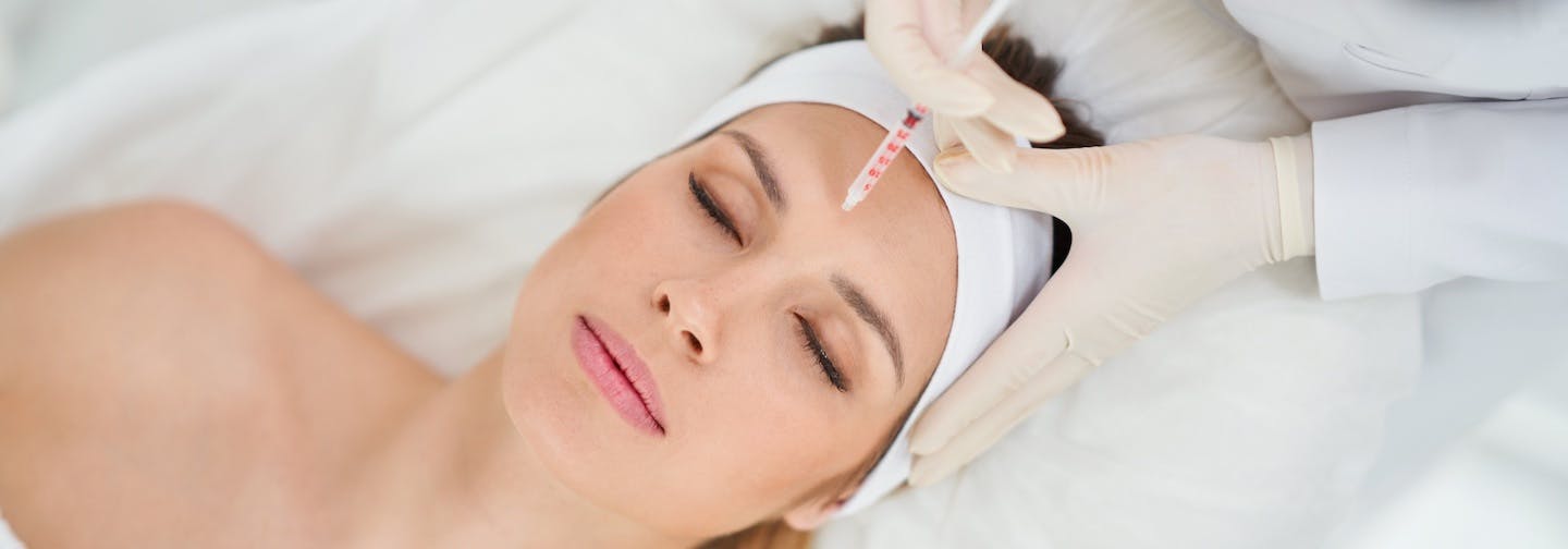 aerial view, gloved hands hold neuromodulator near woman's forehead