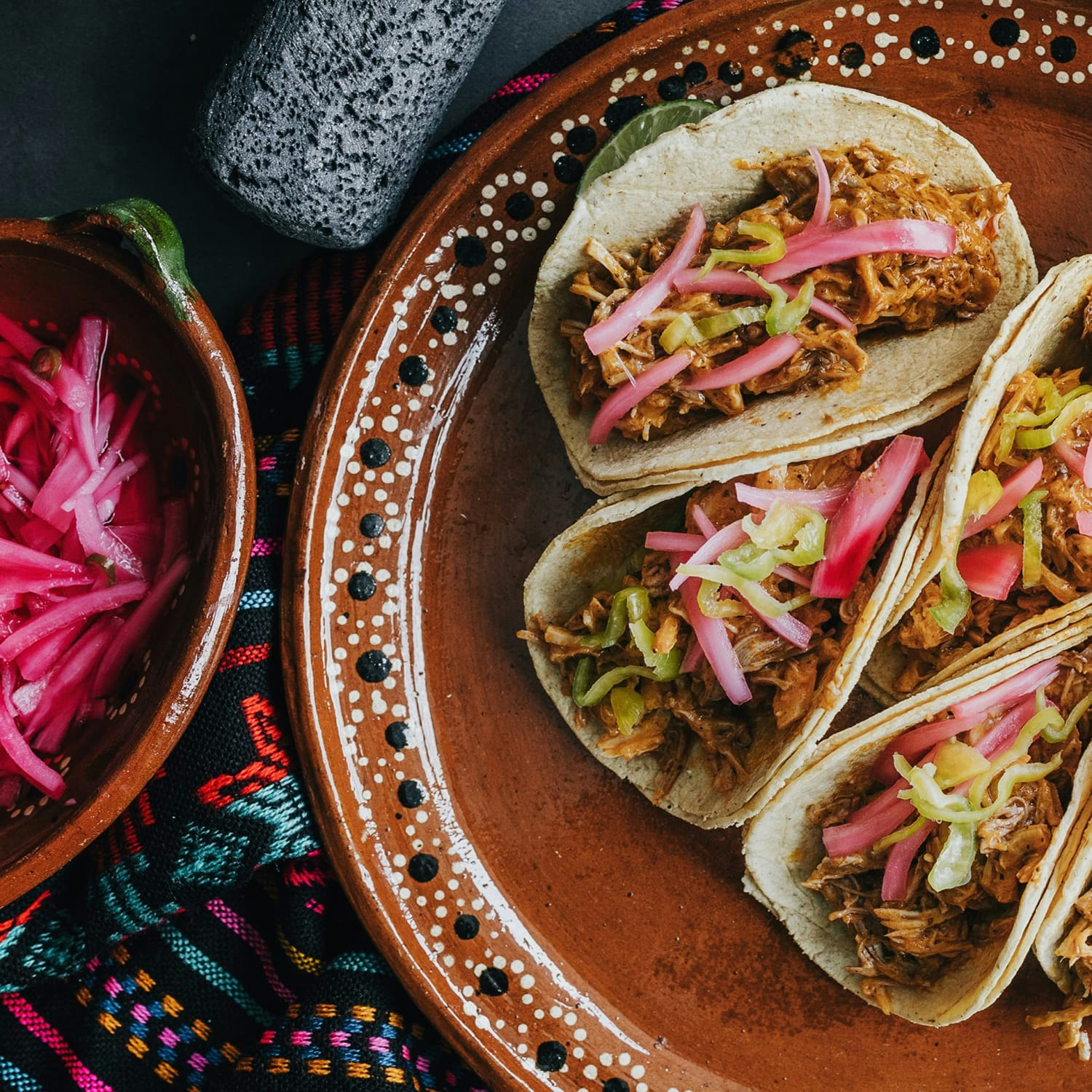 Tacos on a brown plate, sitting on a colorful embroidered tablecloth.