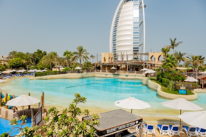 Scenic view of Wild Wadi Waterpark with lounge chairs, umbrellas, and a calm pool, framed by palm trees and the Burj Al Arab standing tall in the background.
