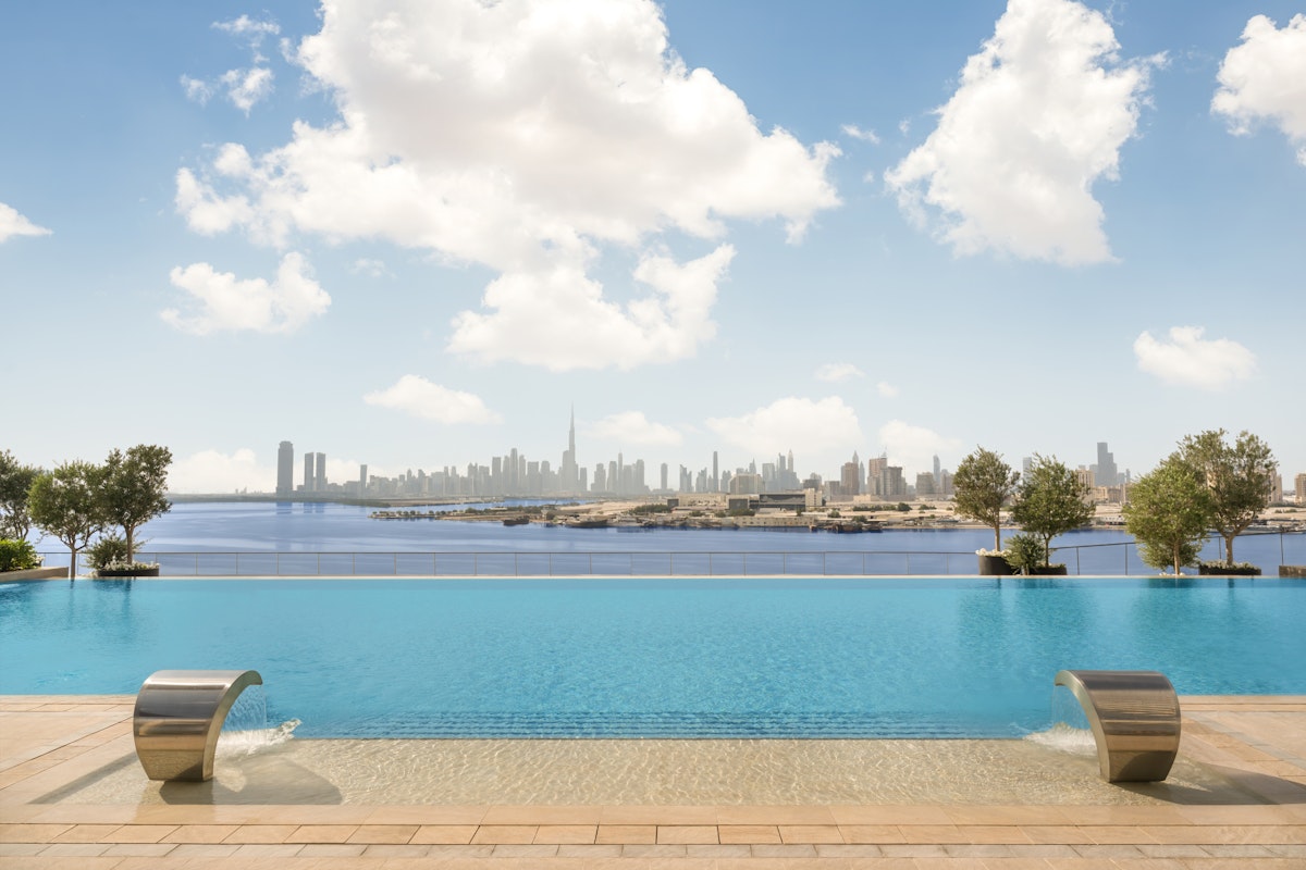 Infinity pool with a view of a distant city skyline under a bright blue sky with scattered clouds, featuring two stainless steel water fountains at the pool's edge