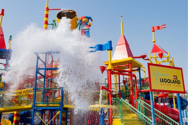 Large splash of water pouring from a giant bucket onto colorful play structures at LEGOLAND Water Park, Dubai, with red, blue, and yellow slides and towers under a sunny sky.