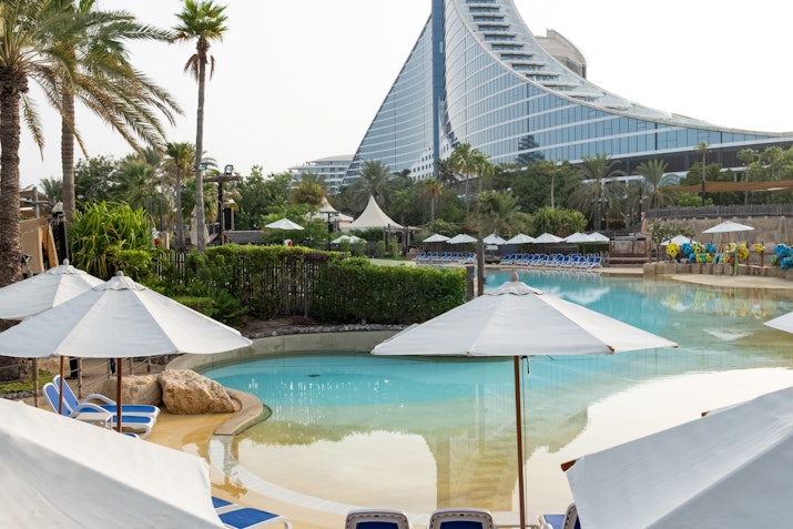 A peaceful lagoon-style pool with white parasols and blue lounge chairs, surrounded by palm trees and greenery, with a large, modern curved building in the background