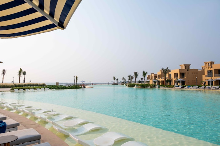 Spacious infinity pool with in-water loungers and waterfront villas in the background under a clear sky