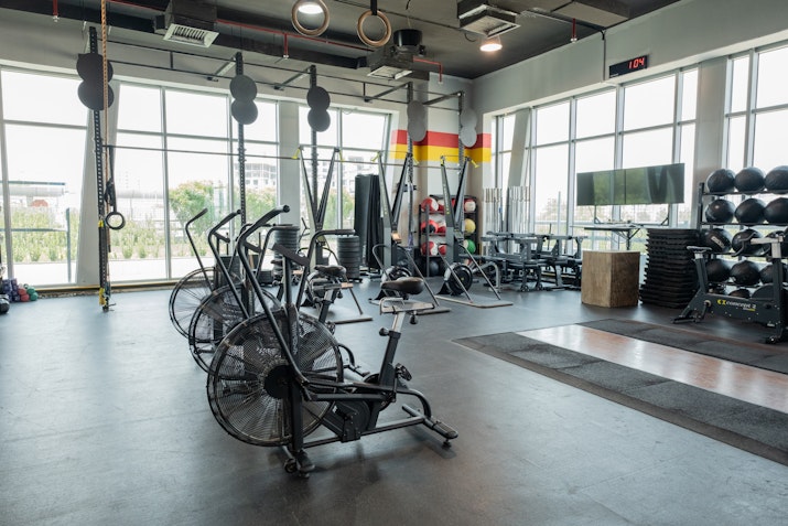 Spacious gym with large windows, featuring air bikes, CrossFit rings, and various workout equipment such as medicine balls, kettlebells, and resistance bands