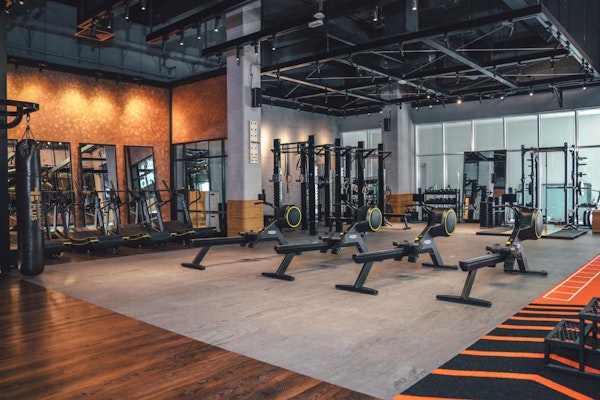 Spacious gym area with rowing machines, a punching bag, wall-mounted fitness equipment, large mirrors, and racks with weights. Industrial-style design with open ceilings and large windows bringing in natural light.