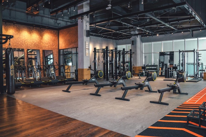 Spacious gym area with rowing machines, a punching bag, wall-mounted fitness equipment, large mirrors, and racks with weights. Industrial-style design with open ceilings and large windows bringing in natural light.