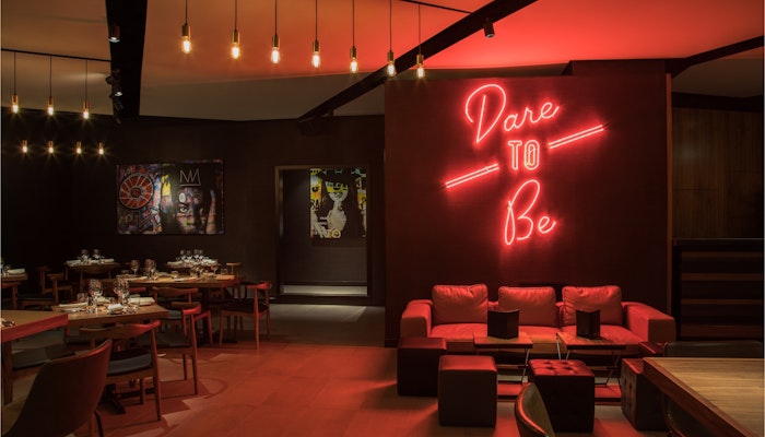 STK Dubai JBR A stylish restaurant interior with dim lighting, featuring a red neon sign that reads 'Dare to Be' above cozy seating, creating an inviting and edgy atmosphere.