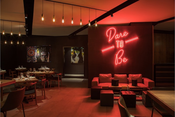 A stylish restaurant interior with dim lighting, featuring a red neon sign that reads 'Dare to Be' above cozy seating, creating an inviting and edgy atmosphere.