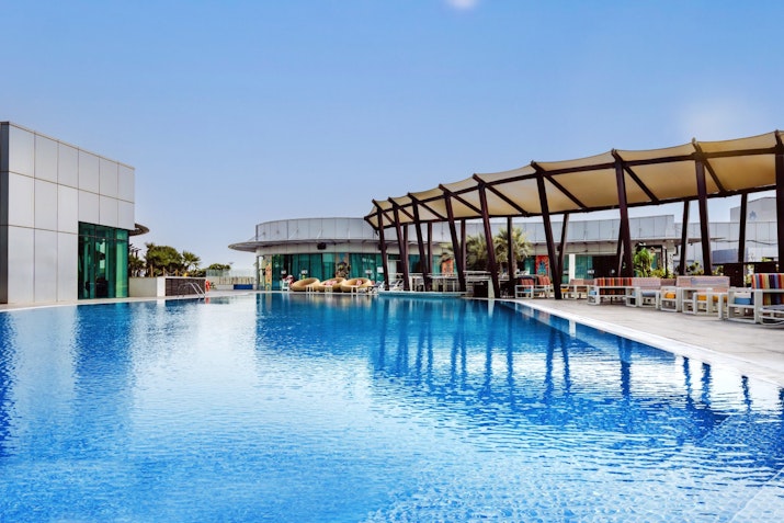 Spacious rooftop pool with clear blue water, surrounded by contemporary buildings and shaded seating areas under a bright sky.