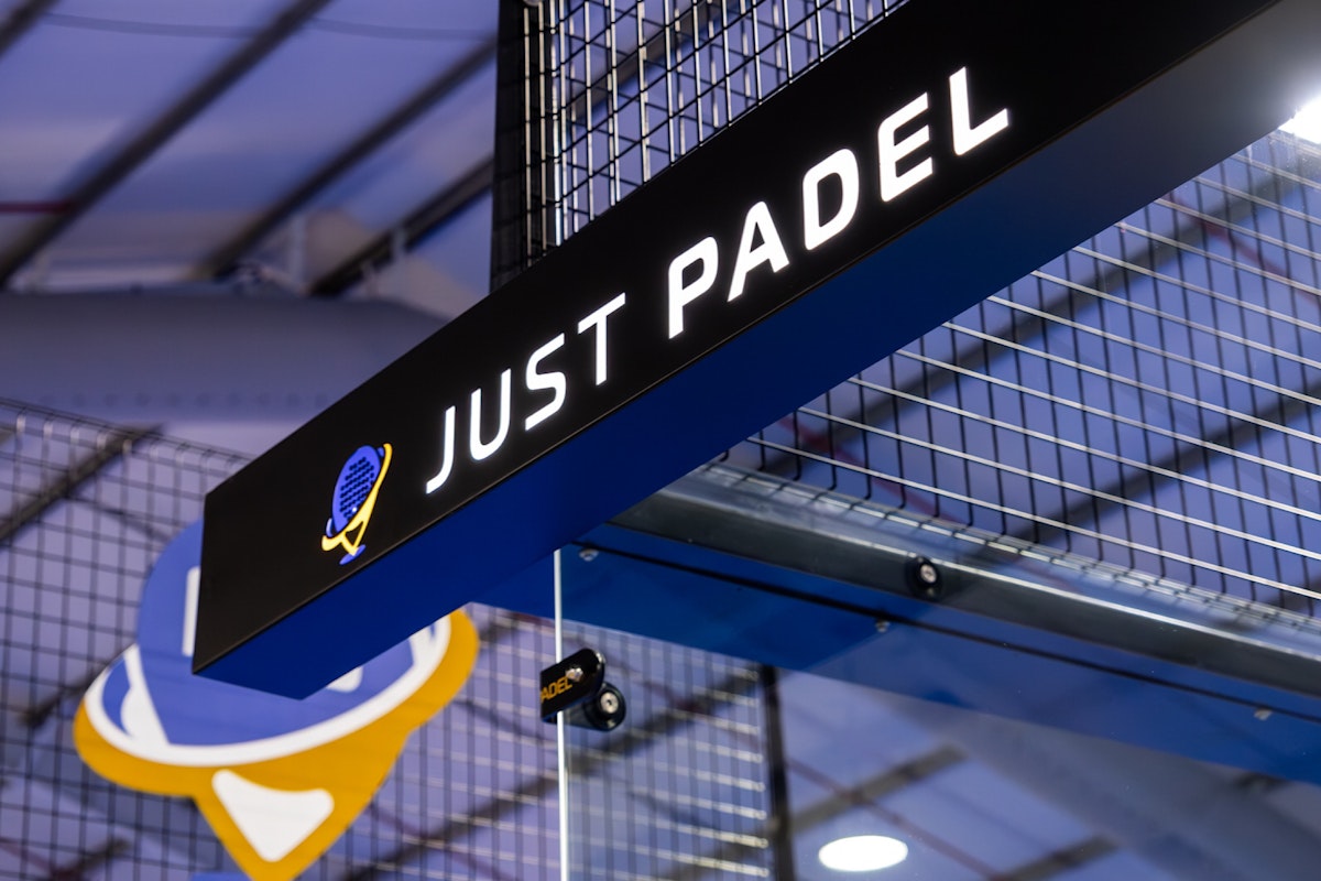 Close-up of the 'Just Padel' sign above a modern indoor padel court, featuring a sleek logo and surrounded by metal fencing, under soft indoor lighting