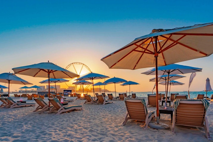 Wavebreaker, Hilton Jumeirah Dubai Sunset view of a beach with rows of wooden loungers and umbrellas on the sand.