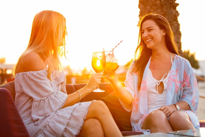 Bliss Lounge, Sheraton Jumeirah Beach Resort Two women enjoying drinks and smiling at sunset, seated outdoors.