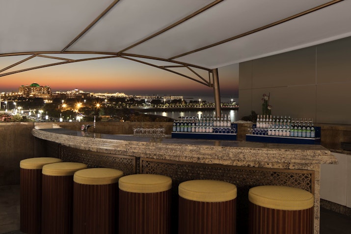 Azura Panoramic Lounge, The St. Regis Elegant rooftop bar with a cityscape view at dusk, featuring high stools and a stone counter.