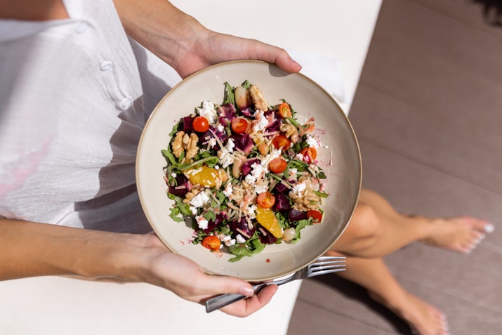 A person holding a bowl of colorful salad with various vegetables and toppings, with their feet visible in the background.