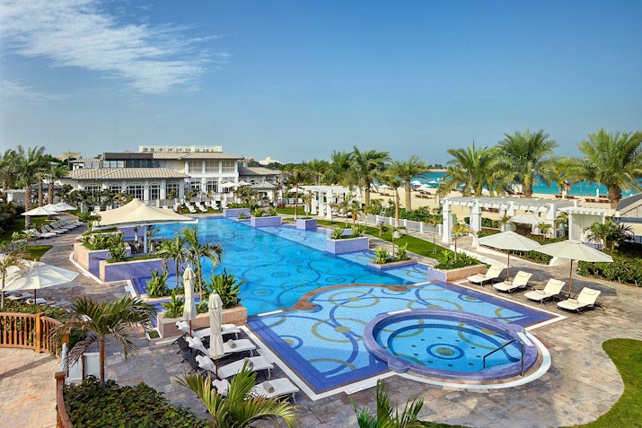 The St. Regis Abu Dhabi Corniche View of a resort pool area with sunbeds, surrounded by palm trees, and a beach in the background.
