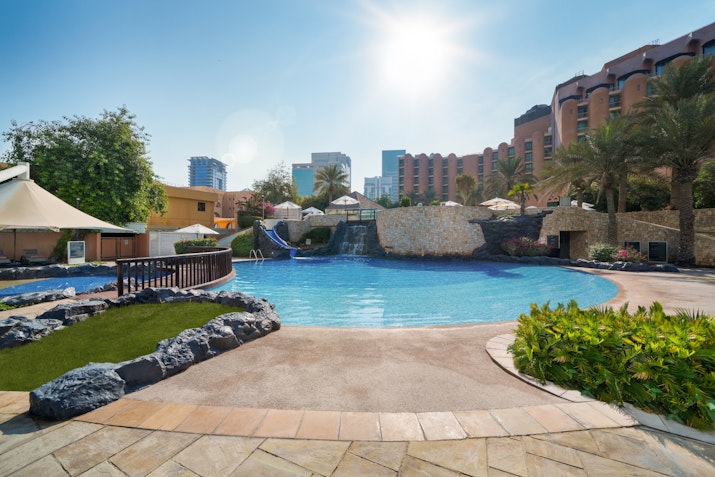 A bright resort pool area with clear blue water, surrounded by lush greenery and rock features, creating a relaxing and inviting atmosphere, under a sunny sky.