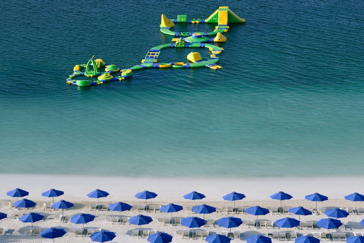 An aerial view of a beach featuring colorful inflatable water attractions in the ocean and rows of blue umbrellas on the sandy shore.