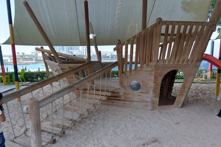 Children's wooden pirate ship playground structure, partially shaded, with a sandy ground and water view in the background.