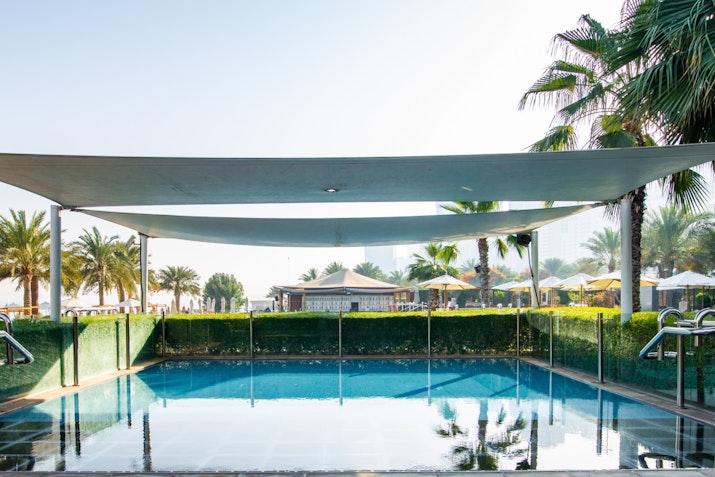 Swimming pool under shaded canopy with palm trees and lounge areas visible in the background
