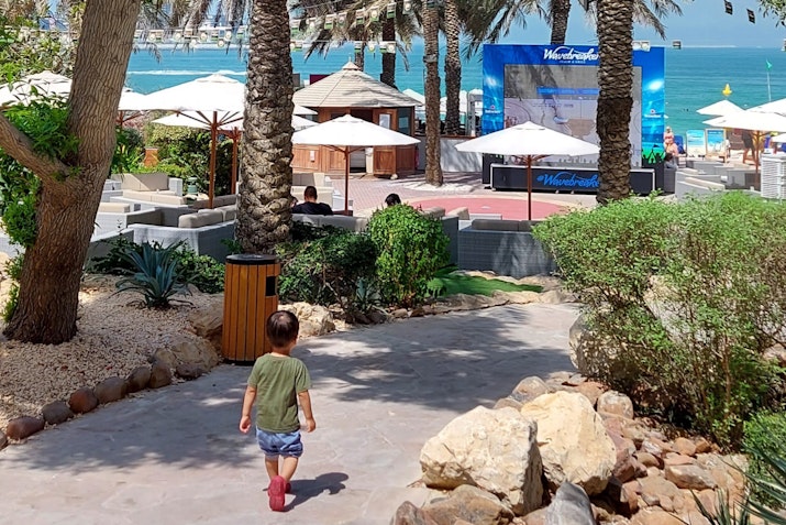A child walking on a pathway near the beach, surrounded by palm trees and umbrellas.