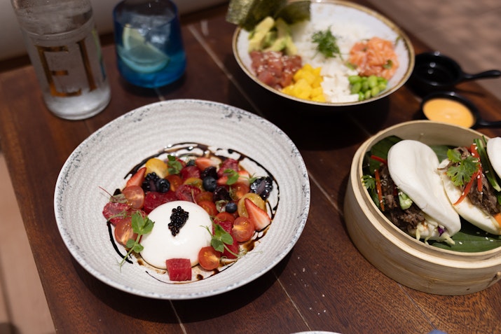 A table setting with a dessert bowl of mixed fruit and yogurt, a bowl of poke with rice and vegetables, and a bamboo steamer filled with bao buns.