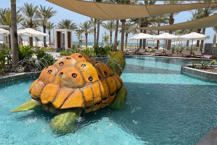 A large turtle sculpture in a swimming pool with palm trees and lounge chairs in the background